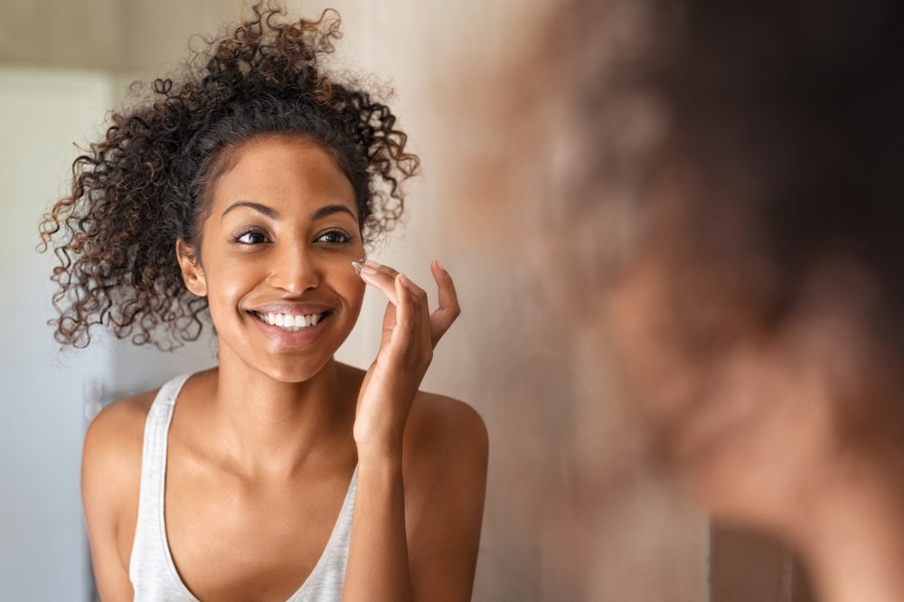 Young woman applying moisturising cream on cheek while standing in front of the mirror in bathroom.